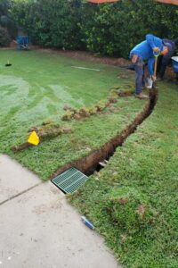 A landscaper trenching a green lawn next to a concrete driveway to install a catch basin and yard drainage system, demonstrating professional lawn care and top garden landscaping in Charlotte, NC.