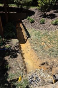 A yard drainage trench excavation featuring a catch basin grate and corrugated pipe, prepared for a French drain system by a lawn care and top garden landscaping service in Charlotte, NC.