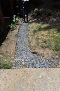 A French drain installation in progress featuring a gravel-filled trench and catch basin grate, completed by a professional lawn care and top garden landscaping service in Charlotte, NC.