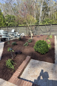 Freshly installed dark landscape mulch in a backyard garden bed featuring low-voltage lighting, shrubs, a brick border, and a stone paver patio in Charlotte, NC